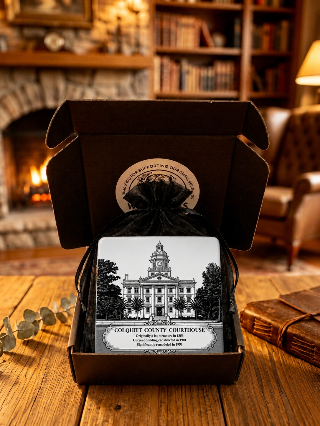 Historic Colquitt County Courthouse ceramic coaster presented in premium gift packaging with a black organza pouch and decorative gift box, displayed on a rustic wooden table in a warm fireside setting.