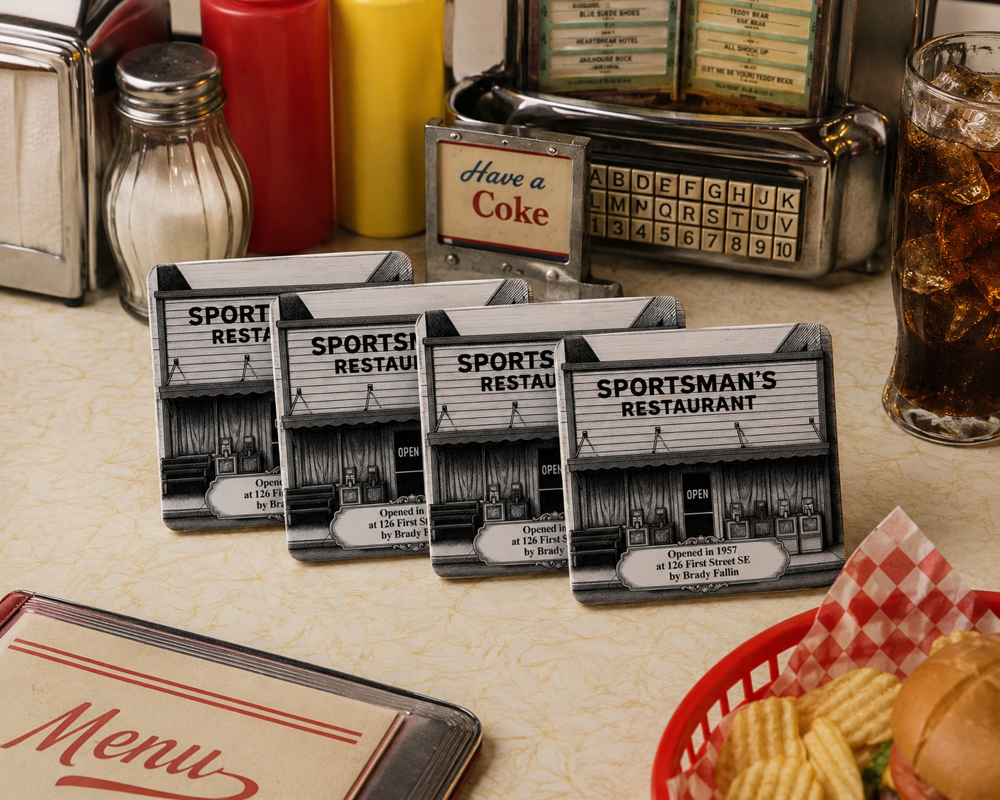 Set of four white ceramic coasters fanned out on a retro diner counter. Coasters feature a black pen-and-ink engraving of Sportsman's Restaurant. Surrounded by a vintage tabletop jukebox, a glass of cola, a menu, and a basket with a burger.