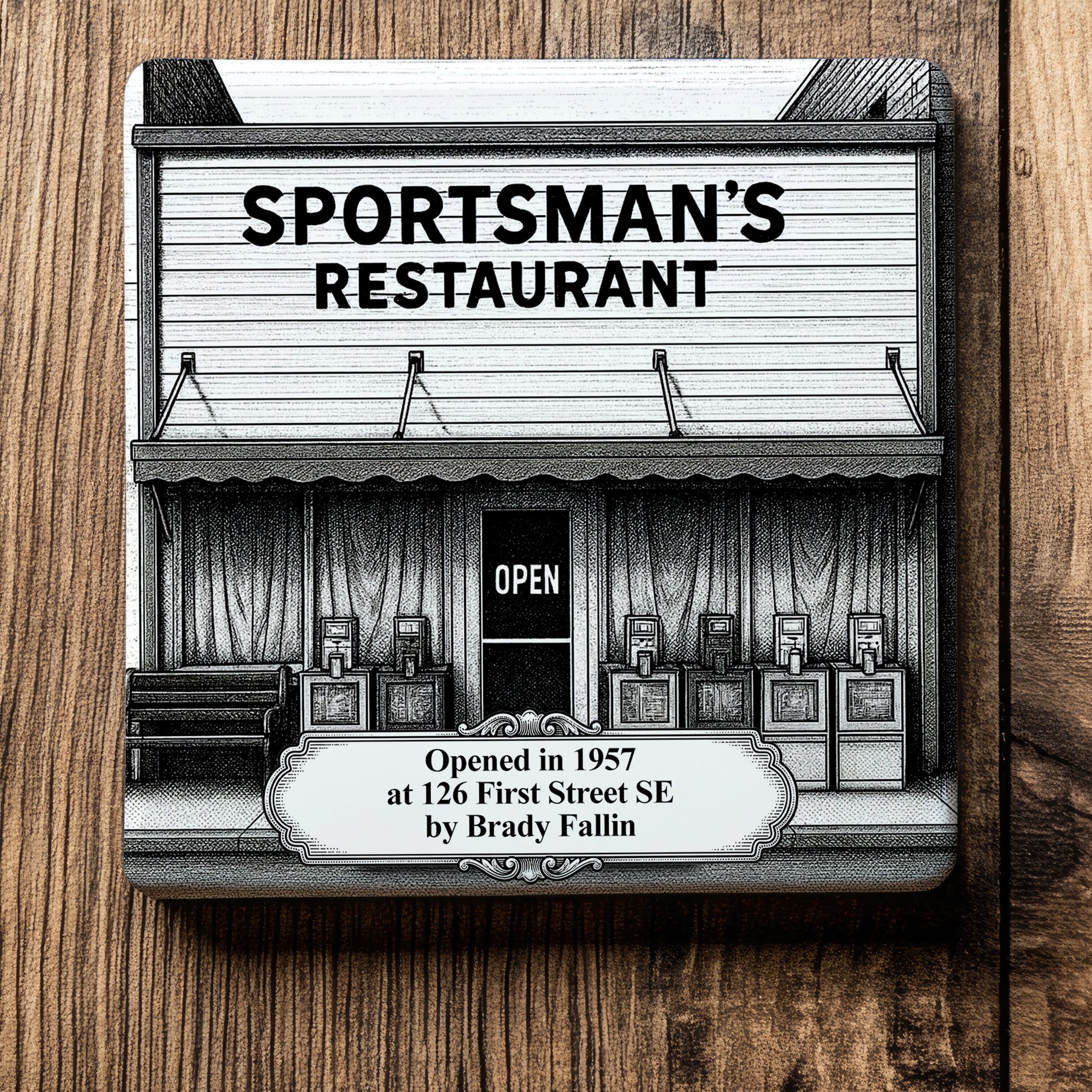 White ceramic coaster with a vintage pen-and-ink engraving of Sportsman's Restaurant in Moultrie, Georgia. Depicts the historic building storefront, an "OPEN" sign, and an ornate timeline text banner, displayed flat on a wood grain background.