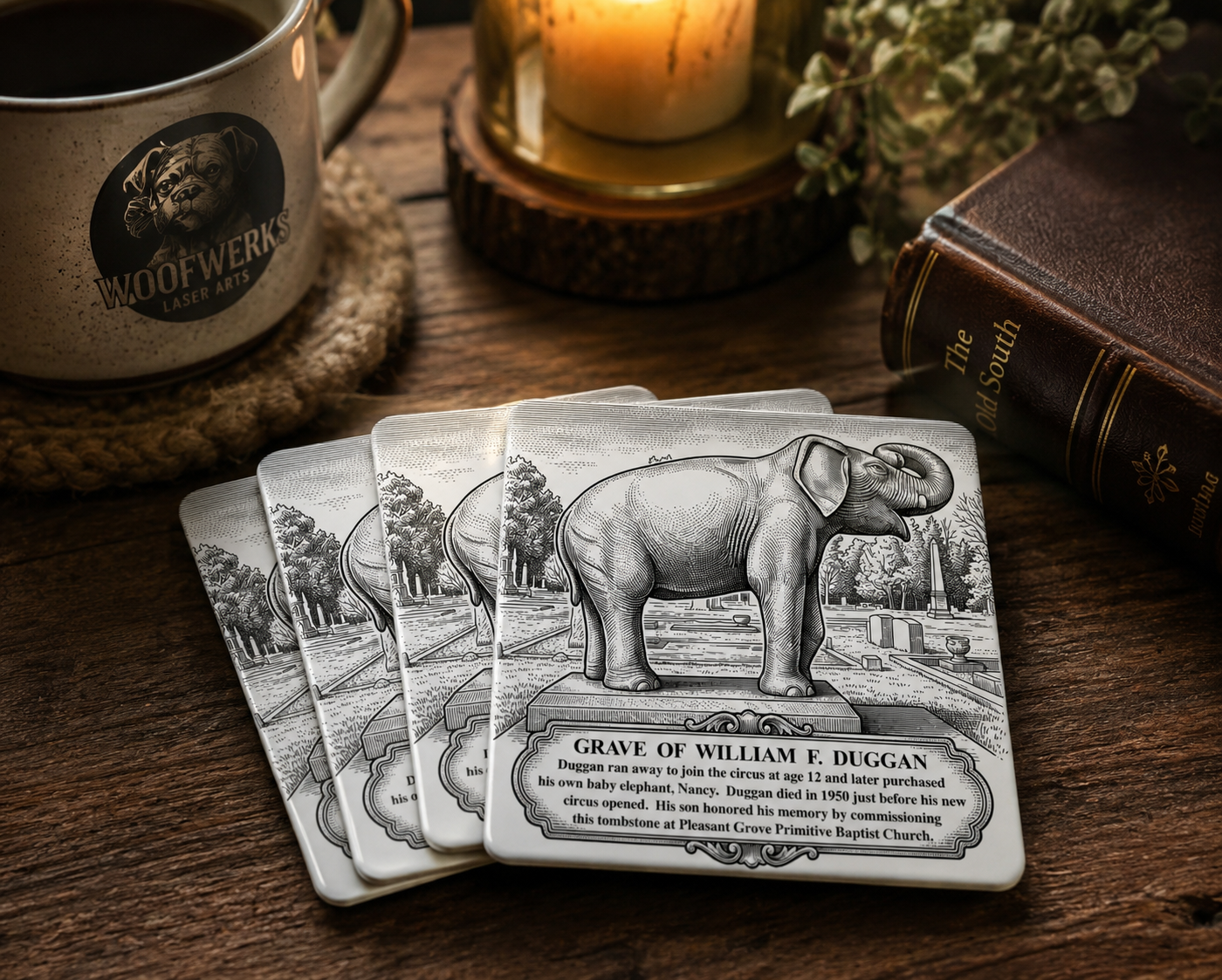 Set of four William F. Duggan Elephant Grave commemorative ceramic coasters displayed on a rustic wooden table with a coffee mug, candle, and vintage book. Part of the Historic Moultrie Coaster Collection.